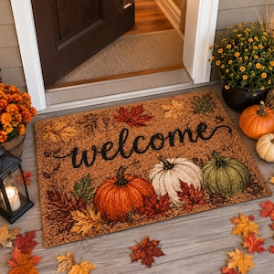 May include: A rectangular coir doormat featuring the word welcome in black script, surrounded by a printed design of orange, white, and green pumpkins, along with scattered autumn leaves in shades of red, yellow, and brown on a tan background.