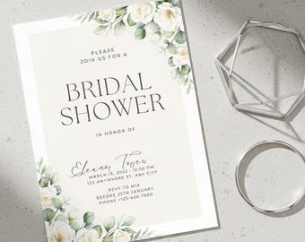 May include: A bridal shower invitation with floral accents and the text "Bridal Shower." The invitation is accompanied by a silver geometric object and a silver ring. Greenery and white flowers decorate the invitation.
