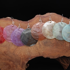 Puede incluir: Un conjunto de siete pendientes redondos en varios colores: rojo, rosa, morado, gris, marrón, beige y azul. Los pendientes están colgados de ganchos plateados y se exhiben sobre un trozo de madera con un fondo oscuro. Un conjunto de joyas colorido.