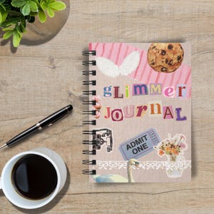 May include: A spiral-bound journal with the words "glimmer journal" in colorful cut-out letters. The cover features a collage of images, including a cookie, angel wings, and a vintage ticket. A black pen and a cup of coffee sit beside the journal.