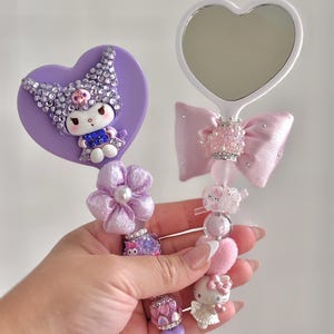 May include: Two decorative hand mirrors. One is heart-shaped with a white frame and a pink bow. The other is purple with a cartoon character. Both mirrors have decorative beaded handles with charms and embellishments.