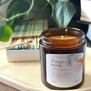 Op de afbeelding: Een bruine glazen potkaars met het label "Pumpkin Patch" met de geurnoten pompoen, kaneel en dennen. De kaars staat op een houten dienblad, met een doosje lucifers en groen in de achtergrond. De kaars is 100 g.
