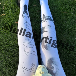 Może przedstawiać: Białe rajstopy z czarnymi wzorami graficznymi i tekstem. Jedna noga ma sylwetkę i napis "This Is My Design". Druga noga ma wzór pszczoły i oka. Rajstopy są noszone z czarnymi butami na trawiastym podłożu.