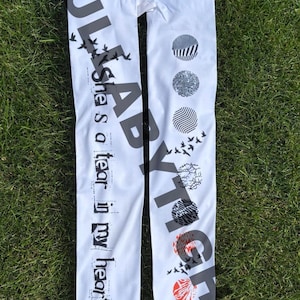 May include: White leggings with black text and graphic designs. The text reads "She is a tear in my heart" and "ABARYXTS". The leggings feature various circular patterns and flying birds.