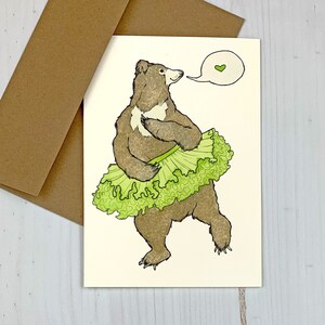 May include: A brown bear wearing a green lettuce tutu and a speech bubble with a green heart. The card is blank inside.