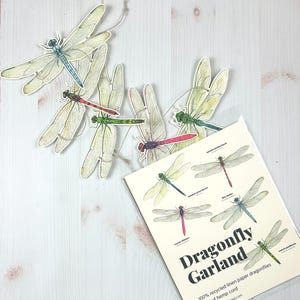 May include: A dragonfly garland with 10 different dragonfly designs printed on recycled linen paper. The garland is 5 feet long and is strung on hemp cord. The text on the card reads "Dragonfly Garland 100% recycled linen paper dragonflies on 5' of hemp cord www.sarahdawithieng.com"