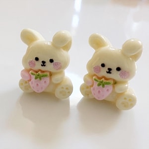 May include: A pair of yellow bunny rabbit earrings with pink and green strawberry accents. The bunnies are holding the strawberries in their paws.