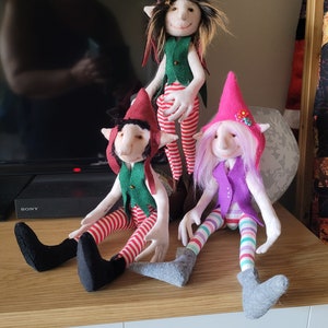 May include: Three handmade felt elf dolls with colorful striped pants and hats. The elf on the left is wearing a green vest, the elf in the middle is wearing a green vest, and the elf on the right is wearing a purple vest.