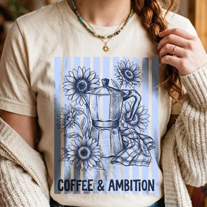 May include: A cream-colored short-sleeve t-shirt featuring a blue graphic print of a moka pot surrounded by sunflowers and a checkered cloth. The design includes vertical blue stripes and the text COFFEE & AMBITION in a bold, dark blue font.