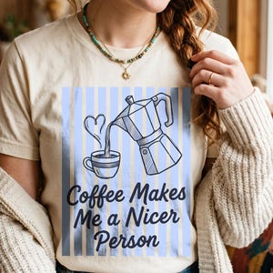 May include: A cream-colored t-shirt featuring a graphic of a moka pot pouring coffee into a cup with a heart-shaped steam swirl. The design is set against a background of vertical light blue stripes and includes the text Coffee Makes Me a Nicer Person.