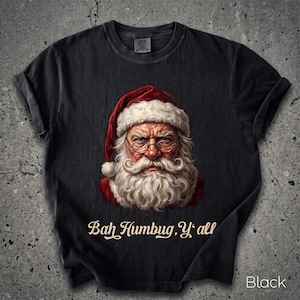 Könnte beinhalten: Ein schwarzes Kurzarm-T-Shirt mit einer Grafik eines mürrischen Weihnachtsmanns, der eine runde Brille und eine rote Mütze trägt. Unter der Abbildung steht der Text "Bah Humbug, Y'all" in einer cremefarbenen Schreibschrift. Rundhalsausschnitt.