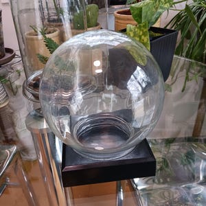 Peut inclure: Un dôme en verre transparent avec une base noire posé sur une table en verre. Le dôme est vide et semble être destiné à la présentation.