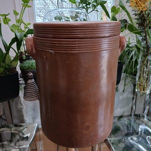 May include: A brown ceramic crock with a lid, sitting on a metal stand. The crock has a textured surface and is slightly weathered.
