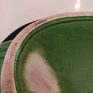 May include: Close-up of the bottom of a green ceramic pot with a pink rim. The pot is marked "JAPAN" on the bottom.
