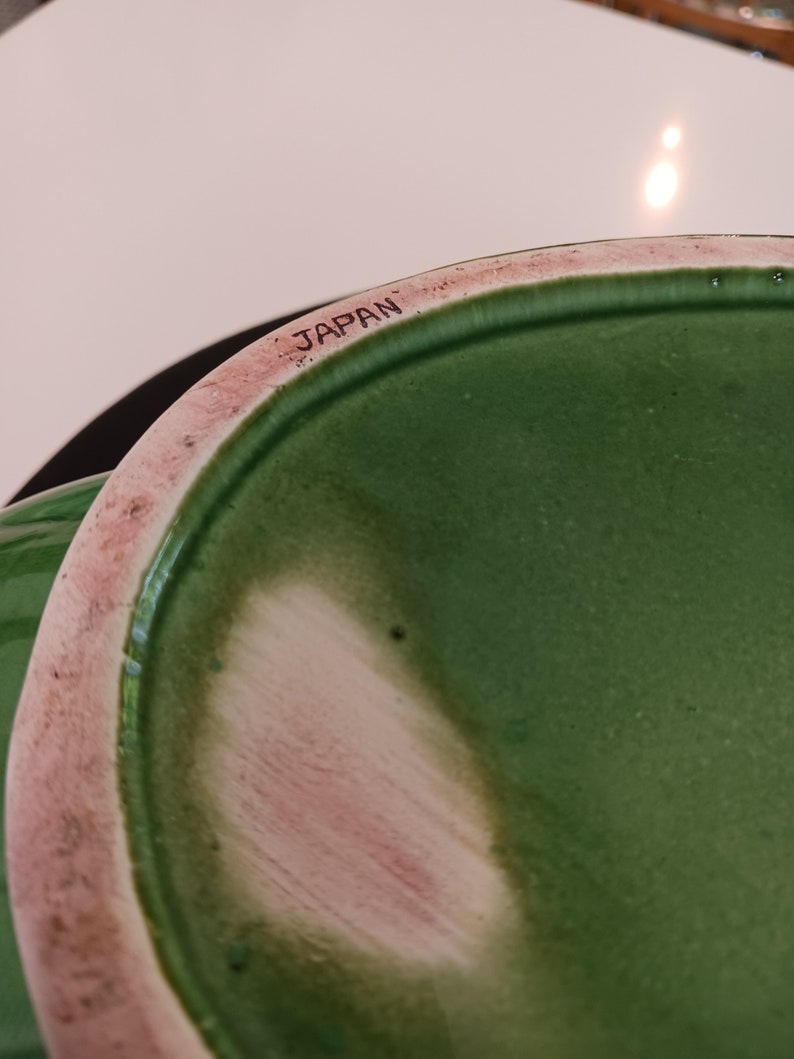 May include: Close-up of the bottom of a green ceramic pot with a pink rim. The pot is marked "JAPAN" on the bottom.