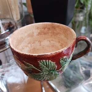 May include: A brown ceramic teacup with a green leaf design. The cup is empty and has a worn look.