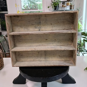 Può includere: Scaffale in legno rustico a tre ripiani con finitura chiara e anticata. I ripiani sono sostenuti da un supporto nero a forma di piedistallo. L'unità è vuota, adatta per esposizione o stoccaggio.