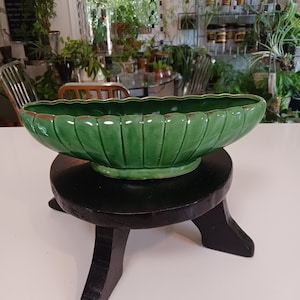 May include: A green ceramic planter with a fluted design sits on a black wooden stand. The planter is shaped like a leaf and has a glossy finish.