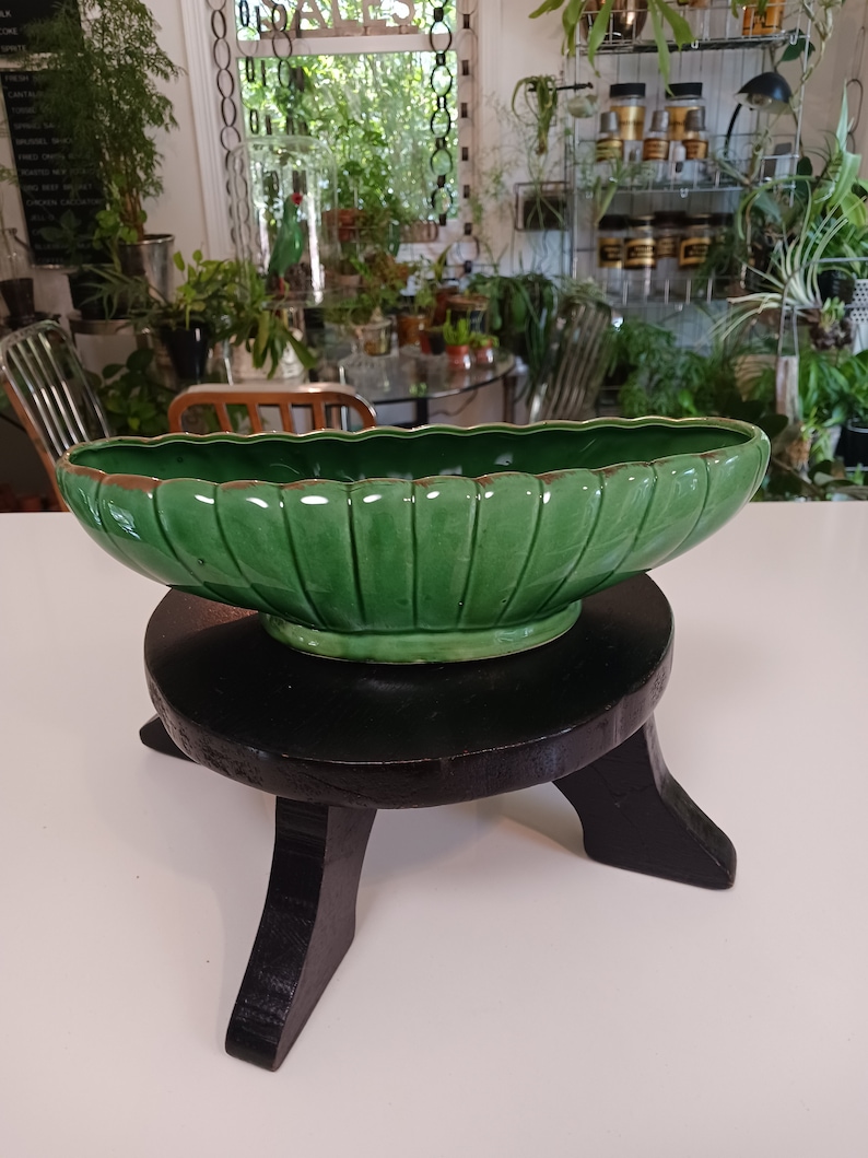 May include: A green ceramic planter with a fluted design sits on a black wooden stand. The planter is shaped like a leaf and has a glossy finish.