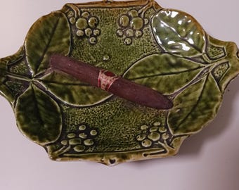 May include: A green ceramic ashtray with a leaf and berry design holds a brown cigar. The ashtray has a glossy finish and a rectangular shape with rounded edges. The cigar has a red and white band.