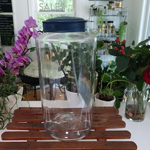 May include: A clear glass storage jar with a cylindrical body and a flat, dark blue metal lid. The jar features a wide mouth and a smooth, transparent surface, providing a clear view of the interior. The lid sits securely on the top rim.