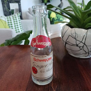 May include: A clear glass Schweppes Ginger Ale bottle with red and white labels. The bottle is on a dark brown wooden surface. The label reads "Schweppes Ginger Ale Dry Non Alcoholic".