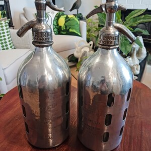 May include: Two vintage soda siphons with metal tops and glass bodies. The siphons have a silver finish and feature a lever-style dispenser. Each bottle has a series of oval cutouts and etched text. The siphons are displayed on a dark wooden table.