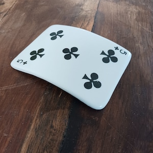 May include: A white, playing card-shaped coaster featuring the design of the five of clubs. The black clubs symbols are evenly spaced across the surface, with a "+5" in the corners. The coaster is set on a dark wood surface.