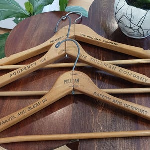 May include: A collection of vintage wooden clothes hangers. Each hanger has a metal hook and is engraved with text, including "PROPERTY OF THE PULLMAN COMPANY" and "TRAVEL AND SLEEP IN". The hangers are arranged on a dark wooden surface.