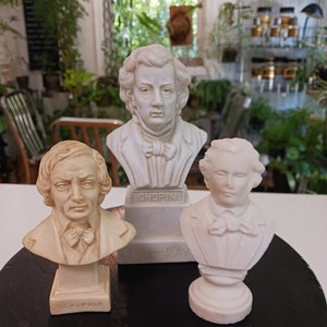 Può includere: Tre busti in gesso bianco di famosi compositori. Il busto a sinistra è etichettato "Schumann", il busto al centro è etichettato "Chopin 1810-1849", e il busto a destra non è etichettato.