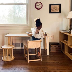 May include: A child's play area with a light wood table, chair, and stool. A red clock and a framed bicycle print hang on the wall. The child is cutting paper with scissors. A wooden storage crate is on the floor.