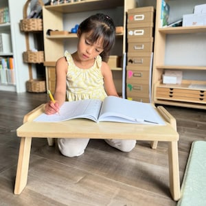 May include: A small, light-colored wooden lap desk with angled legs. The desk is open, displaying a notebook. A young person is seated in front of the desk, writing with a pencil. The person is wearing a yellow floral dress.