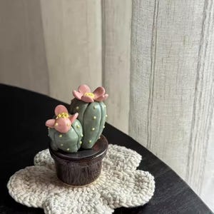May include: A ceramic cactus sculpture with pink flower accents sits in a dark brown pot, placed on a cream-colored, flower-shaped crochet coaster. The cactus is light green with yellow details. The scene is set on a dark table.