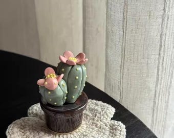May include: A ceramic cactus sculpture with pink flower accents sits in a dark brown pot, placed on a cream-colored, flower-shaped crochet coaster. The cactus is light green with yellow details. The scene is set on a dark table.