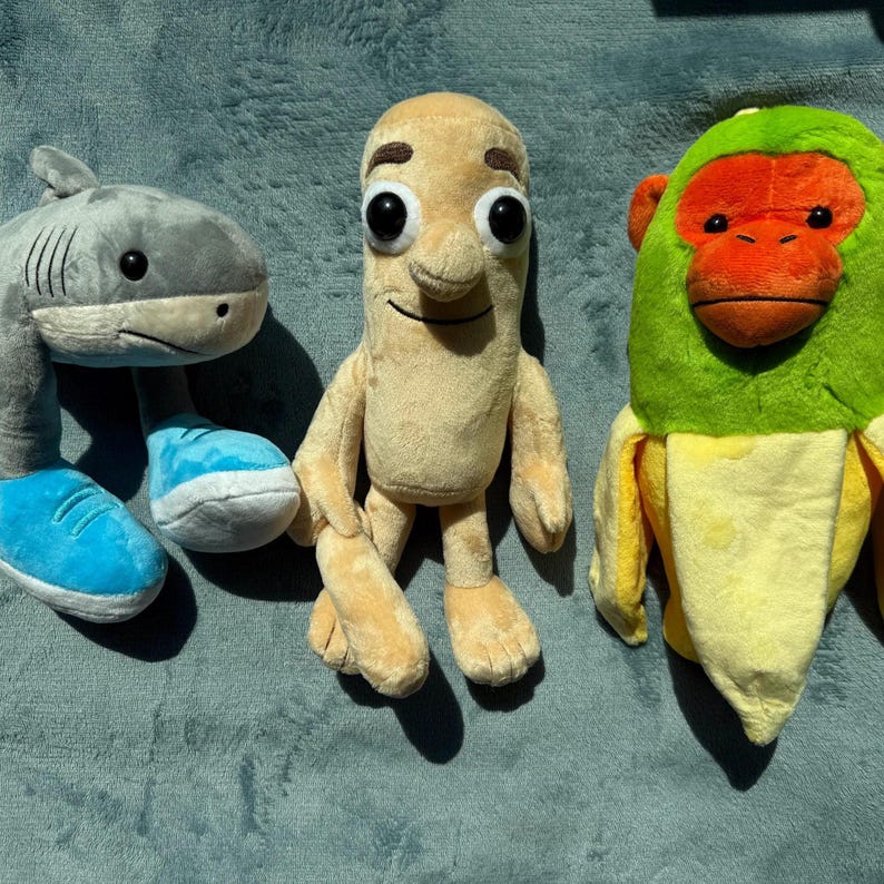 May include: Three plush toys: a gray shark wearing blue sneakers, a tan bean-shaped character, and a monkey in a banana peel. The monkey has a green head and a red face. The toys are on a blue surface.