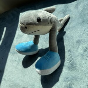 May include: A gray plush shark toy with a white belly, black eyes, and blue sneakers. The shark has a long neck and a fin on its back. The toy is sitting on a blue surface.