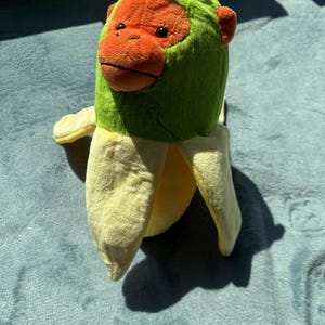 May include: A plush toy featuring a monkey head with an orange face and green hood emerging from a yellow banana peel. The toy is made of soft fabric and is designed to resemble a monkey in a banana.