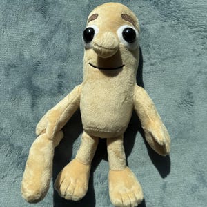 May include: A plush toy in the shape of a light tan, humanoid bean character with large black eyes, a small nose, and a smiling mouth. The toy has arms and feet, and is made of a soft, fuzzy material.