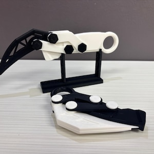 May include: A 3D-printed, black and white claw-shaped object, with a ring handle and articulated segments. One is displayed on a black stand, and another lies flat. The design features geometric shapes and a futuristic aesthetic.