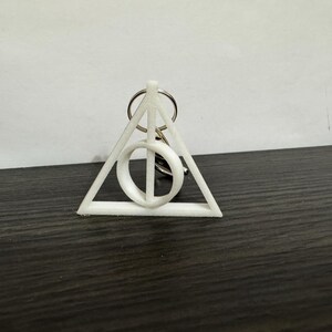 May include: White Deathly Hallows symbol keychain. The keychain features a triangle with a circle inside and a vertical line, all connected to a silver keyring. The symbol is a popular design from the Harry Potter series.