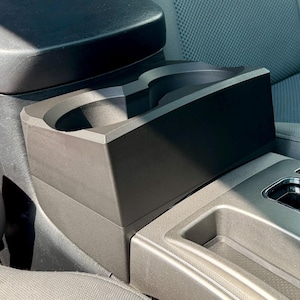 May include: A black car console cup holder with two circular openings, positioned between the front seats. The cup holder is made of a dark material and sits on a gray console. The car interior features gray seats and a gear shift.