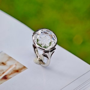 Peut inclure: Une bague en argent avec une grosse pierre précieuse ovale et transparente. La pierre précieuse présente un motif floral gravé. La bague a un anneau décoratif ajouré avec des volutes stylisées. Le fond est vert et blanc.