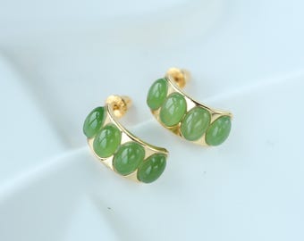 May include: A pair of gold-toned hoop earrings, each adorned with five oval-shaped, light green gemstones. The earrings have a curved design, with the gemstones set along the outer edge. The background is a soft, off-white color.