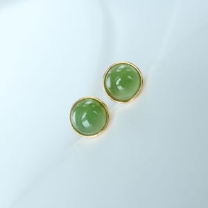 May include: Two round stud earrings with a gold-colored bezel. Each earring features a smooth, translucent, light green stone. The earrings are displayed on a white surface, creating a clean and elegant presentation.