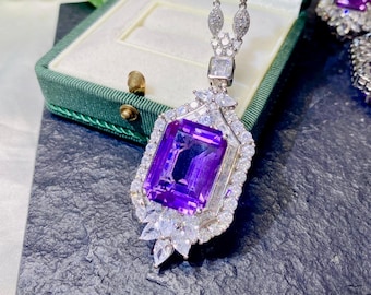 May include: A silver necklace with a large, rectangular, deep purple gemstone pendant. The pendant is surrounded by smaller, clear gemstones. The necklace is displayed in a green jewelry box.