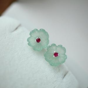 May include: A pair of flower-shaped stud earrings. Each earring has a light green, translucent floral design with a small, round, red gemstone centre. The earrings are set on a white surface.