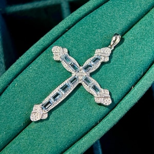 May include: A silver-toned cross pendant featuring a central row of rectangular light blue gemstones. The cross is bordered by small, clear pavé-set stones and has decorative, textured silver-toned metal accents at each of the four ends.
