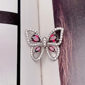 May include: A silver butterfly brooch with a sparkling design. The wings are outlined with small, clear crystals, and the center features heart-shaped and oval-shaped red gemstones. The brooch is a jewelry item.