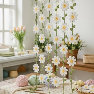 Peut inclure: Un rideau de marguerites crochetées à la main, avec des pétales blancs et des centres jaunes, enfilées sur des tiges vertes. Le rideau est suspendu à une tringle en bois. Des pelotes de laine et des crochets sont sur une table recouverte d'une nappe crème. Un panier de marguerites crochetées est également visible.