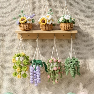 May include: A collection of handmade crochet hanging planters and flower baskets. The planters are filled with colorful crochet flowers and trailing plants. The baskets are suspended from a wooden shelf and macrame hangers. Yarn balls in various colors are at the bottom.